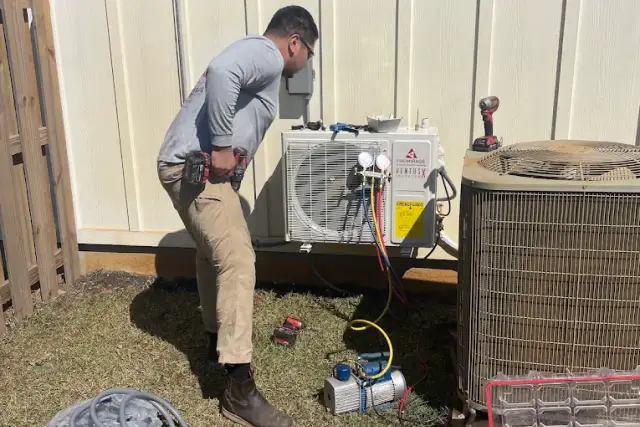 A newly installed ductless mini split condenser being charged with manifold gauges, standing beside the larger existing central air conditioner condenser at the same Texas home