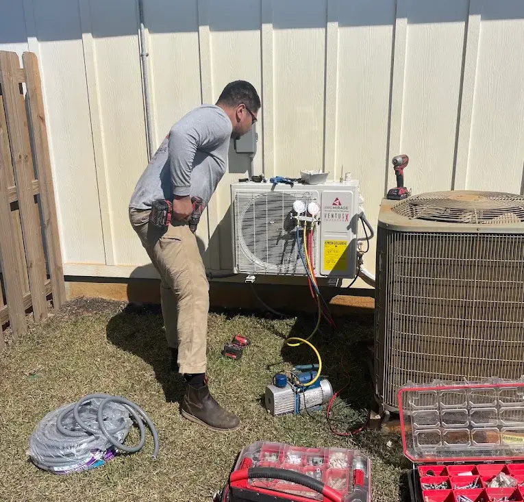A newly installed ductless mini split condenser being charged with manifold gauges, standing beside the larger existing central air conditioner condenser at the same Texas home