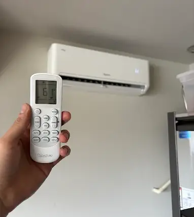 A hand holding a mini split remote control set to 61 degrees in front of a wall-mounted indoor head