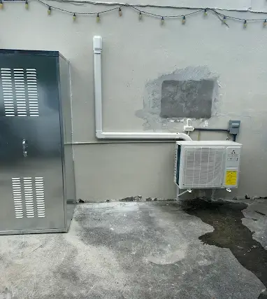 A ductless mini split condenser mounted above wet concrete, with ice and frost still on the wall behind it after a Texas freeze