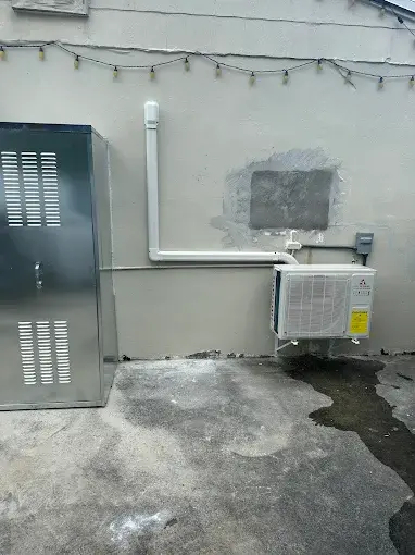 A ductless mini split condenser mounted above wet concrete, with ice and frost still on the wall behind it after a Texas freeze